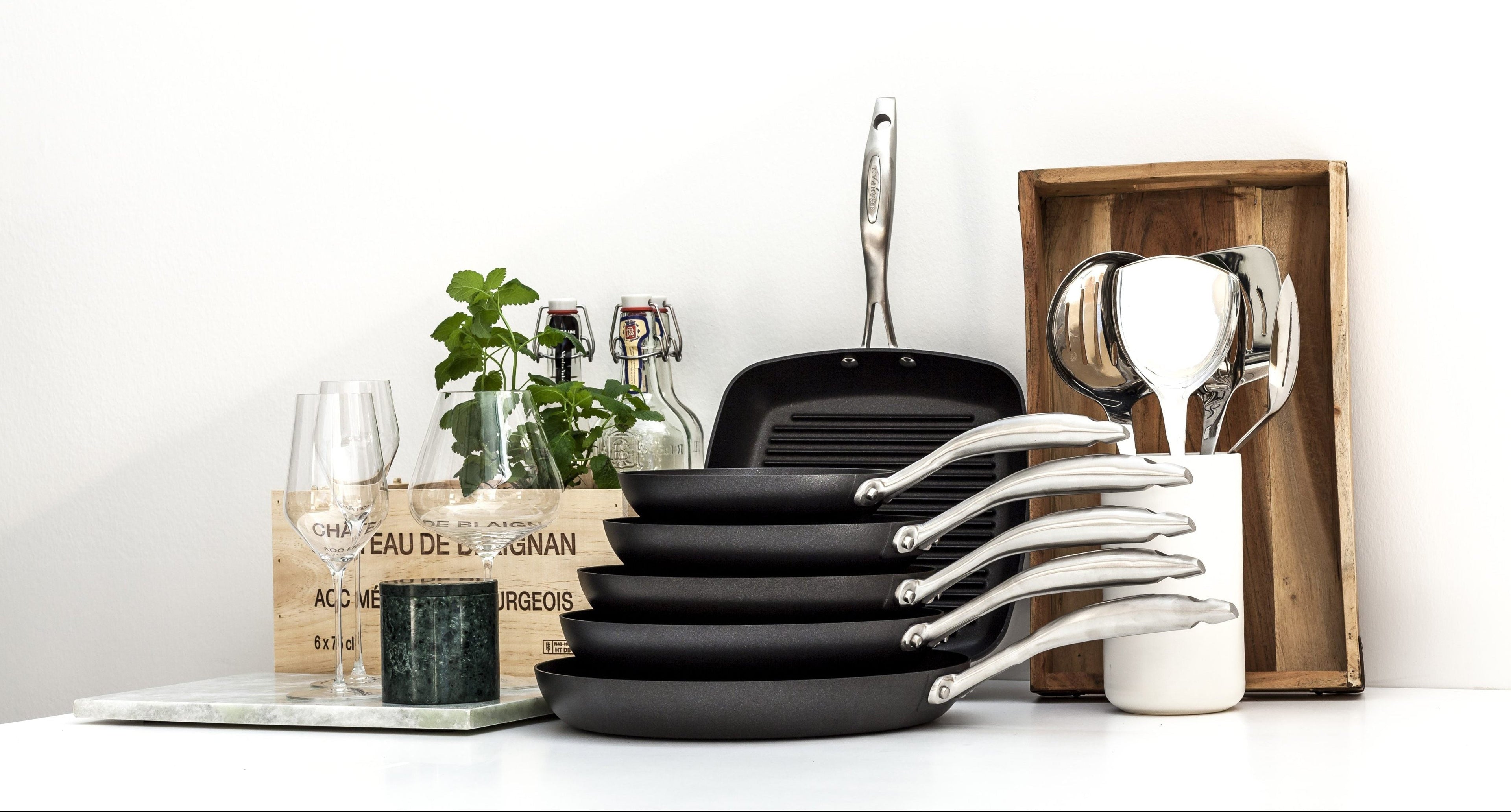 Set of black cookware including pans and spatulas on a white surface with a plant and wooden box in the background.